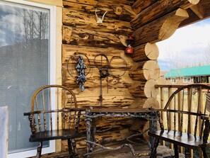 A rustic wooden cabin interior with a wooden table and chairs, a window with blinds, and a view of a green-roofed building outside. - Wrangell Mountain Lodge (Chitina)