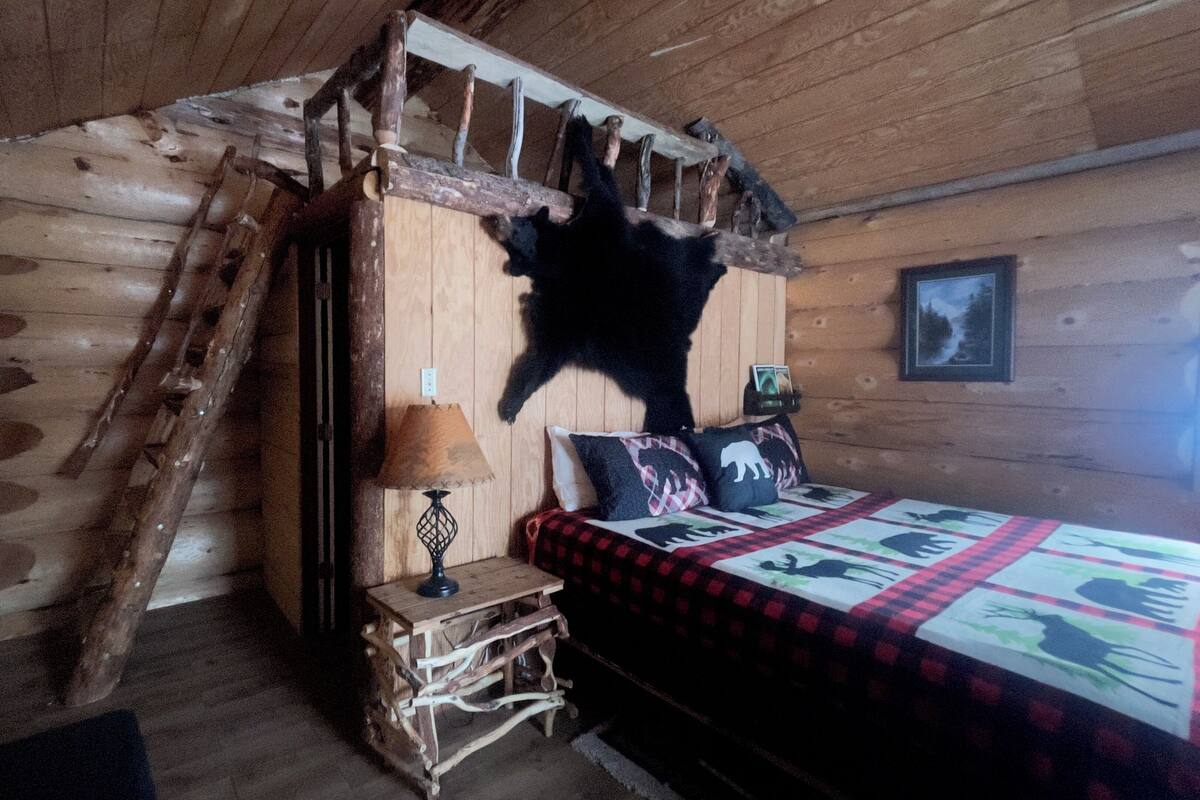Una habitación rústica tipo cabaña con una escalera de madera, una cama con colcha, una lámpara y un trofeo de animal en la pared.