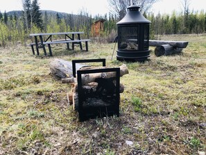 A rustic outdoor setting with a wooden bench, a stone fireplace, and a log structure. - Wrangell Mountain Lodge (Chitina)