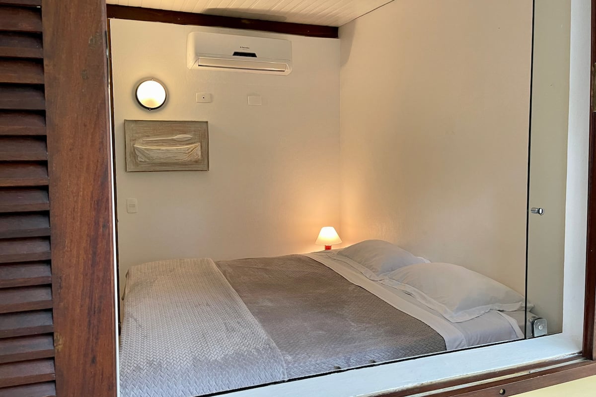 A bedroom with a bed, a bedside lamp, a wall-mounted air conditioner, and a framed picture.