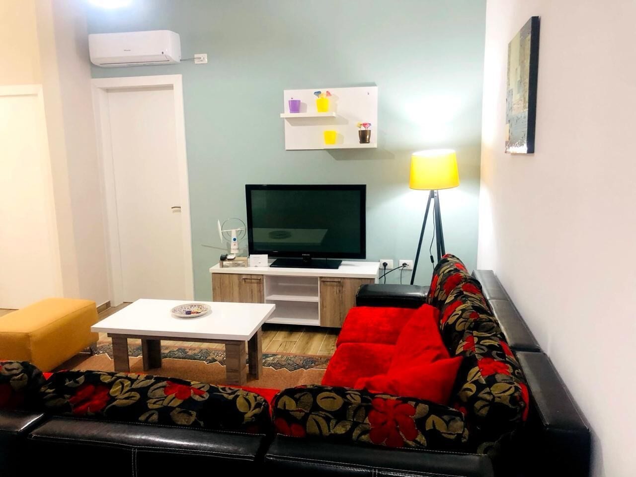 Una sala de estar con sofá, mesa de centro, televisor de pantalla plana, lámpara y estante con objetos decorativos.
