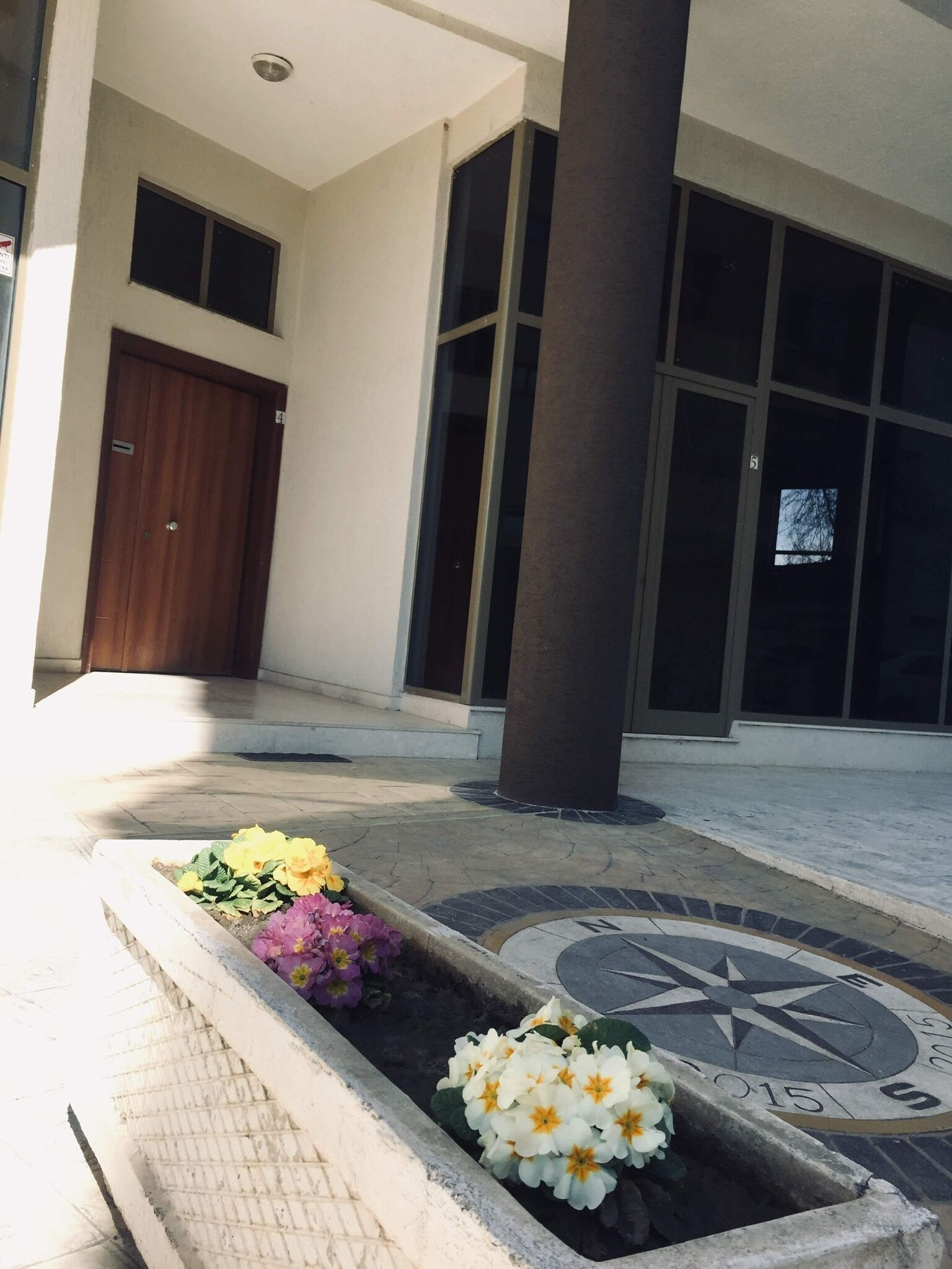 A building entrance with a wooden door, large windows, and a flower bed with various flowers.