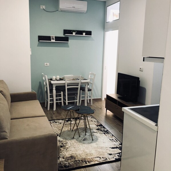 Uma sala de estar com sofá, mesa de jantar com cadeiras, televisão e geladeira.