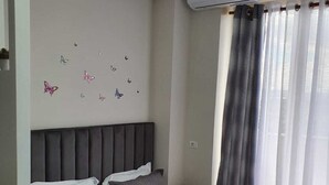 A modern hotel room with a large bed, grey bedding, a headboard, a bedside table, a remote control, and a window with curtains. - Rezidenca Shopi Apartments (Elbasan)