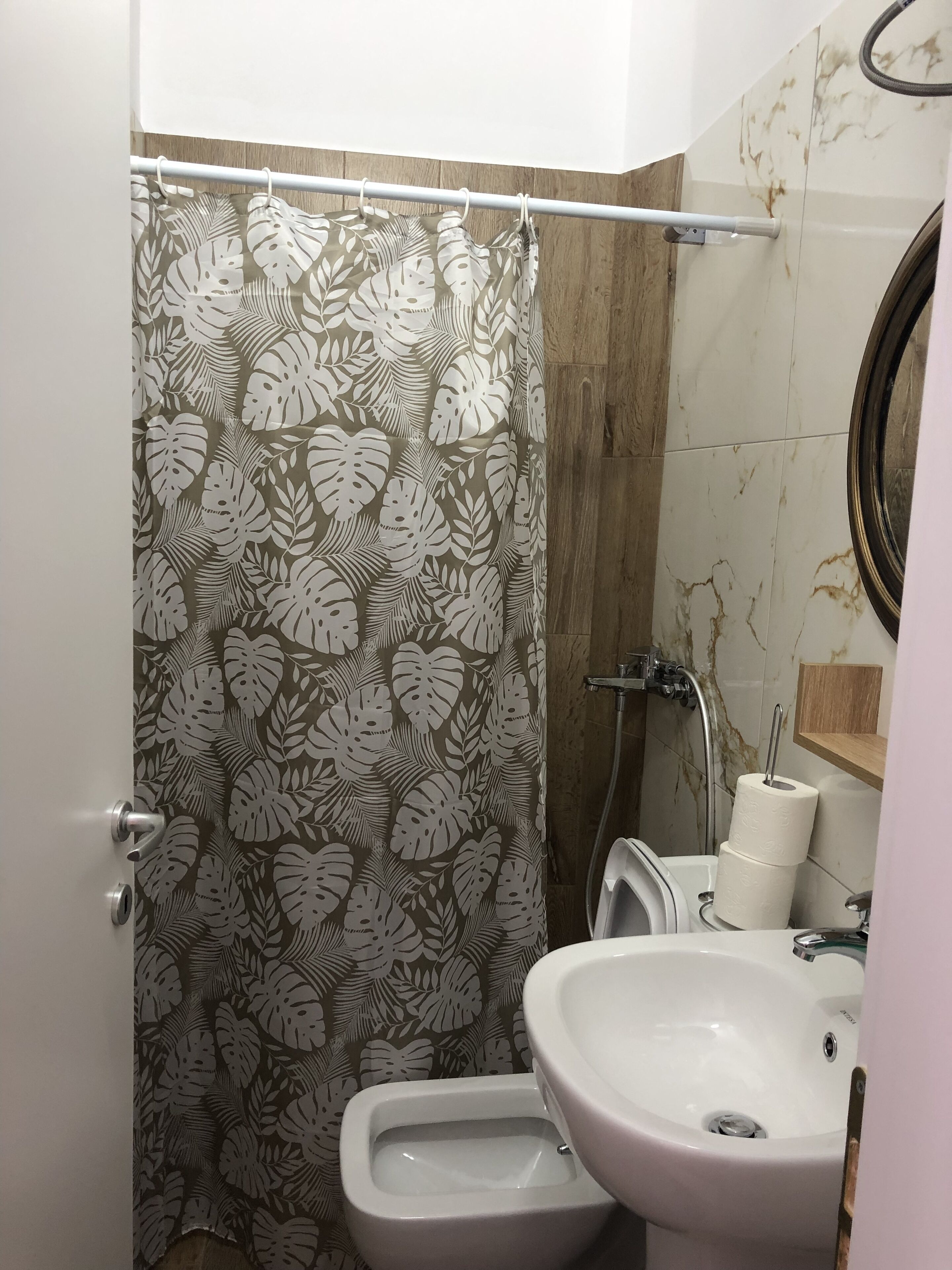 Um banheiro com cortina de chuveiro estampada com folhagens, vaso sanitário, bidê, pia e parede com azulejos de mármore.