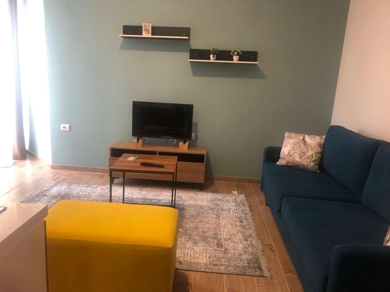 Un salon comprenant un canapé, un pouf jaune, un téléviseur posé sur un meuble en bois et deux étagères ornées d’objets décoratifs.