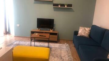 Un salon comprenant un canapé, un pouf jaune, un téléviseur posé sur un meuble en bois et deux étagères ornées d’objets décoratifs.