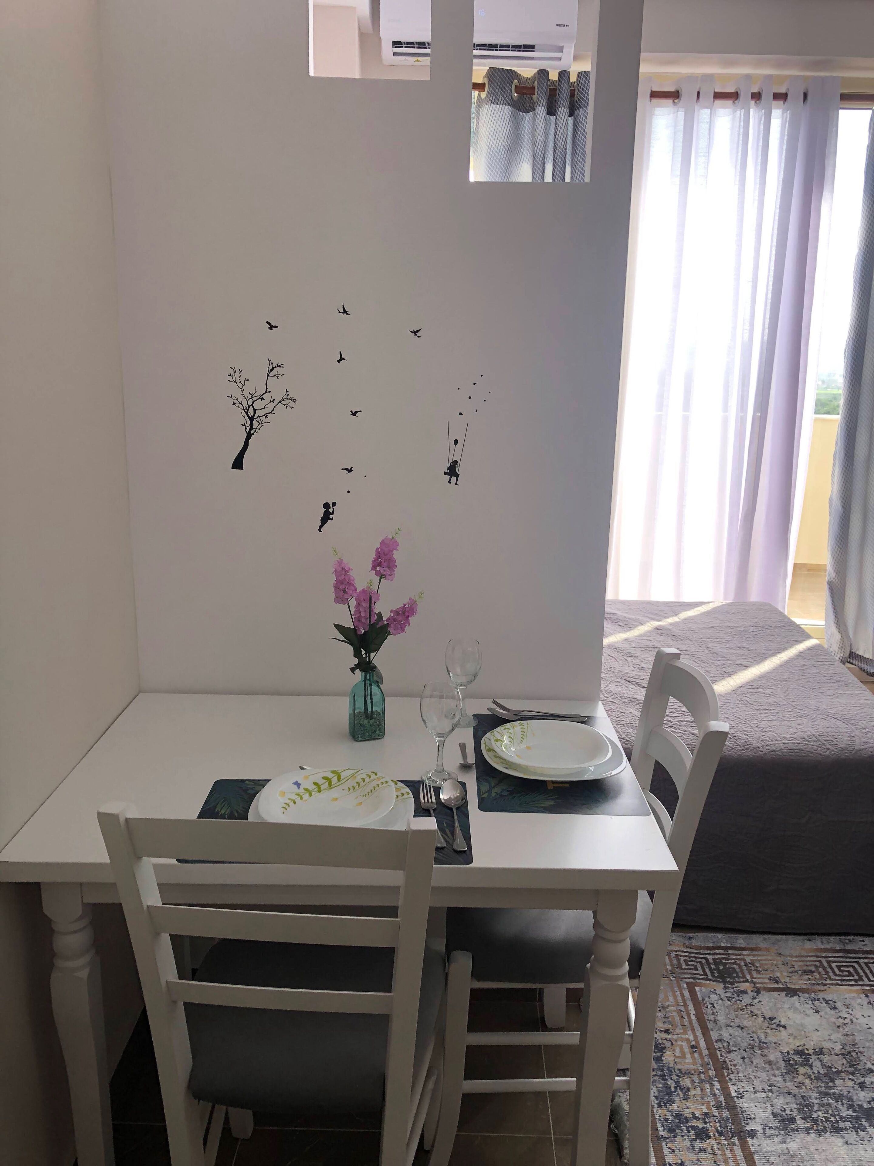 A dining area with a white table, two chairs, a vase with flowers, and wall decals of birds and a tree.
