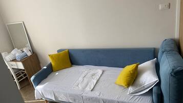 A single bed with white bedding and two yellow pillows, a blue sofa, a wooden desk with a mirror, and a blue chair.