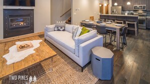 A modern living room with a fireplace, a sofa, a coffee table, and a dining area with a kitchen in the background. - 3BR Big Sky Townhome | Walk to Town Center, Hot Tub & Sleeps 12 (Big Sky)