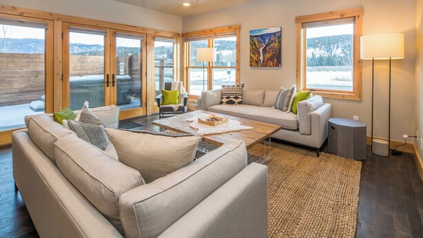 A spacious living room with a large sofa, a coffee table, and a view of a snowy landscape through large windows. - 3BR Big Sky Townhome | Walk to Town Center, Hot Tub & Sleeps 12 (Big Sky)