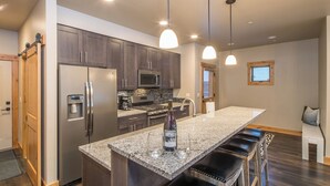 A modern kitchen with dark wood cabinets, stainless steel appliances, a granite countertop, and pendant lighting. - 3BR Big Sky Townhome | Walk to Town Center, Hot Tub & Sleeps 12 (Big Sky)