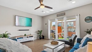 Ein Wohnzimmer mit Flachbildfernseher, Deckenventilator, Couchtisch und Sofa. Ein Schild trägt die Aufschrift 'BEACH HOUSE' und ein weiteres 'ON BEACH TIME'.