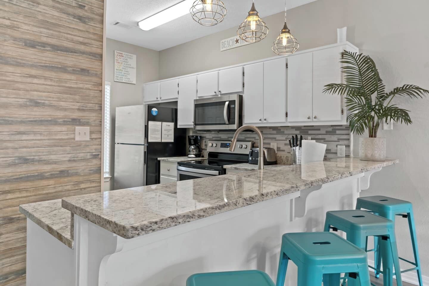 Una cocina moderna con gabinetes blancos, encimeras de granito, electrodomésticos de acero inoxidable y taburetes de bar color turquesa.