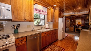 A kitchen with wooden cabinets, a stainless steel dishwasher, a white refrigerator, and a window with a checkered valance. - Fir Forest Cottage by Hauserman Rental Group (Tahoe City)