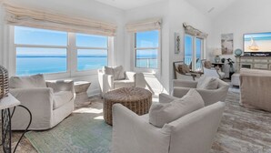 A spacious living room with a view of the sea, featuring a ceiling fan, large windows, and a television. - Condos directly on the Gulf (Destin)