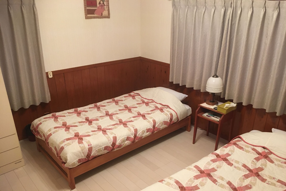 A room with two single beds, wooden headboards, a bedside table with a lamp, and a small nightstand with a book and a tissue box.