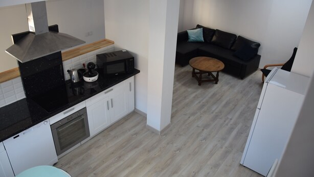 A compact kitchen with a microwave, oven, and sink. There is a black countertop, white cabinets, and a wooden dining table. A black sofa and a white refrigerator are also visible. - The edge of Saint Quentin 10 minutes from METZ at the foot of Mont Saint Quentin (Lessy)