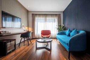 A modern living room with a blue sofa, a red chair, a glass coffee table, a flat-screen TV, and a large window with curtains.