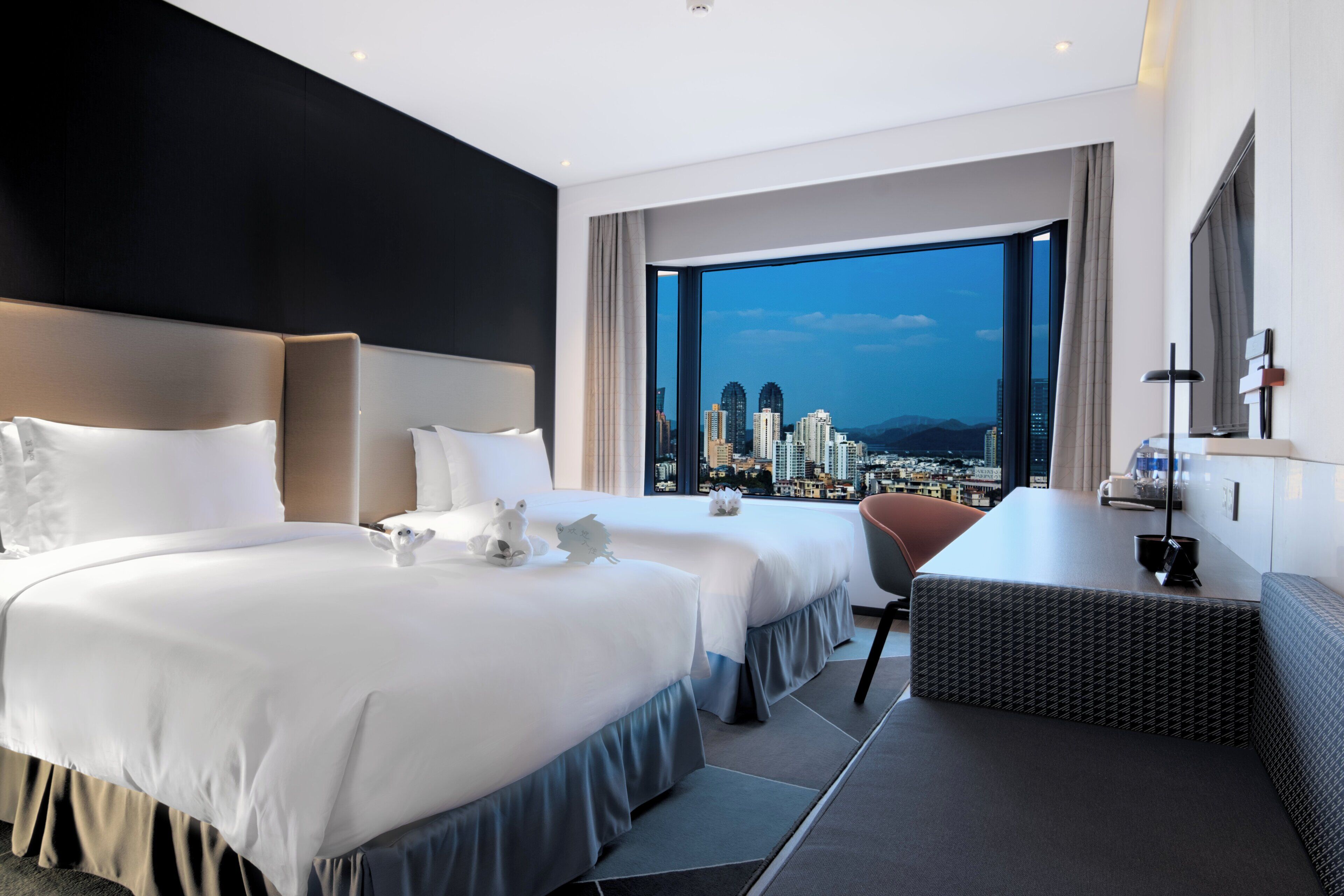 Habitación de hotel con dos camas, un escritorio y un ventanal grande con vistas a la ciudad.