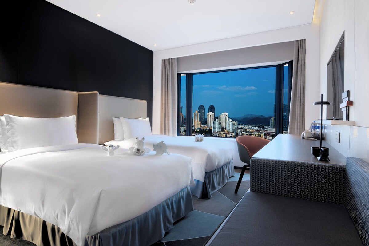 A hotel room with two beds, a desk, and a large window offering a city view.