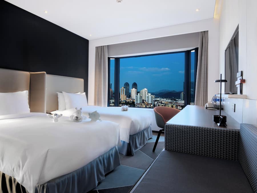 A hotel room with two beds, a desk, and a large window offering a city view.