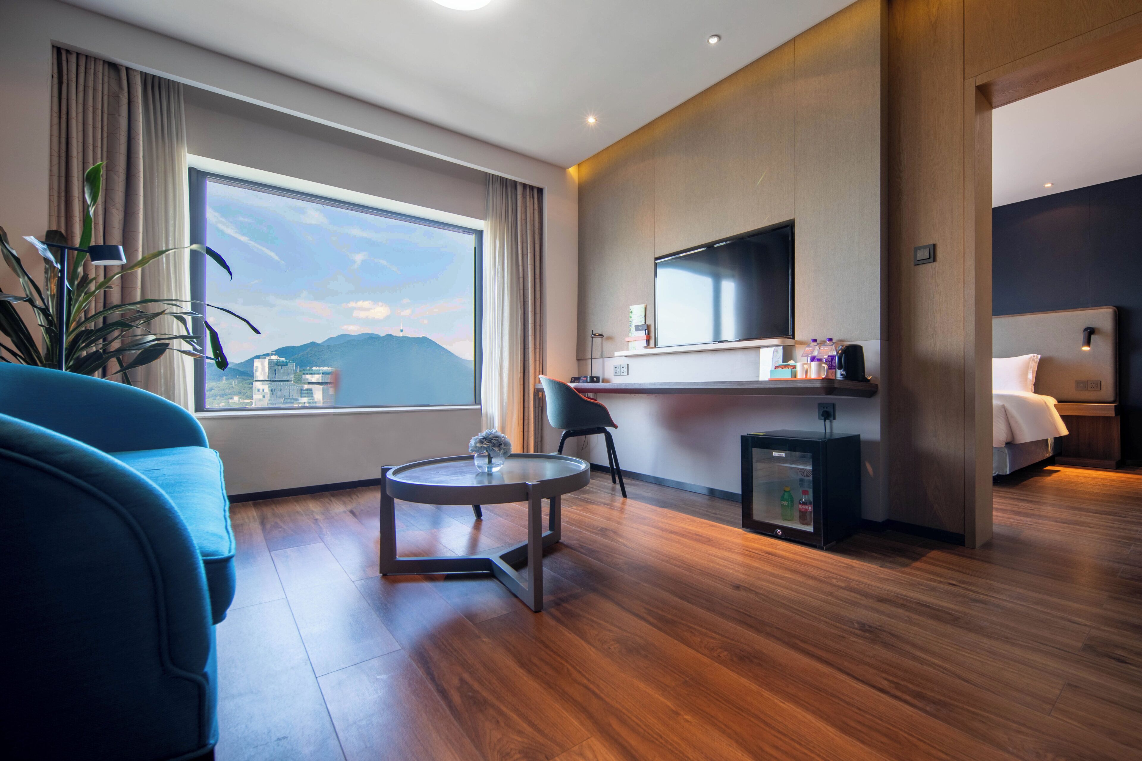 A modern hotel room with a large window, a flat-screen TV, a small table, a chair, and a bed.