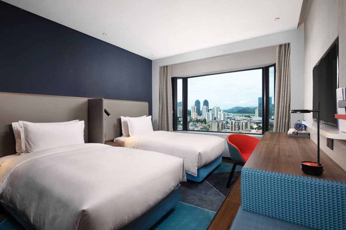 A hotel room with two beds, a desk, a chair, and a large window offering a city view.