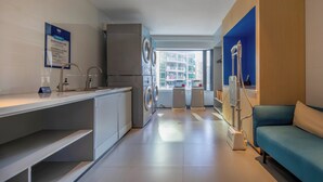 A modern laundry room with a washing machine, dryer, and ironing station. There is a blue sofa and a window providing a view of the city. - Holiday Inn Express Shenzhen Dongmen by IHG (Shenzhen)