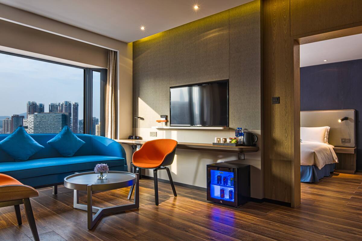 A modern hotel room with a city view, a blue sofa, an orange chair, a small coffee table, a flat-screen TV, a mini-fridge, and a bed with a headboard.