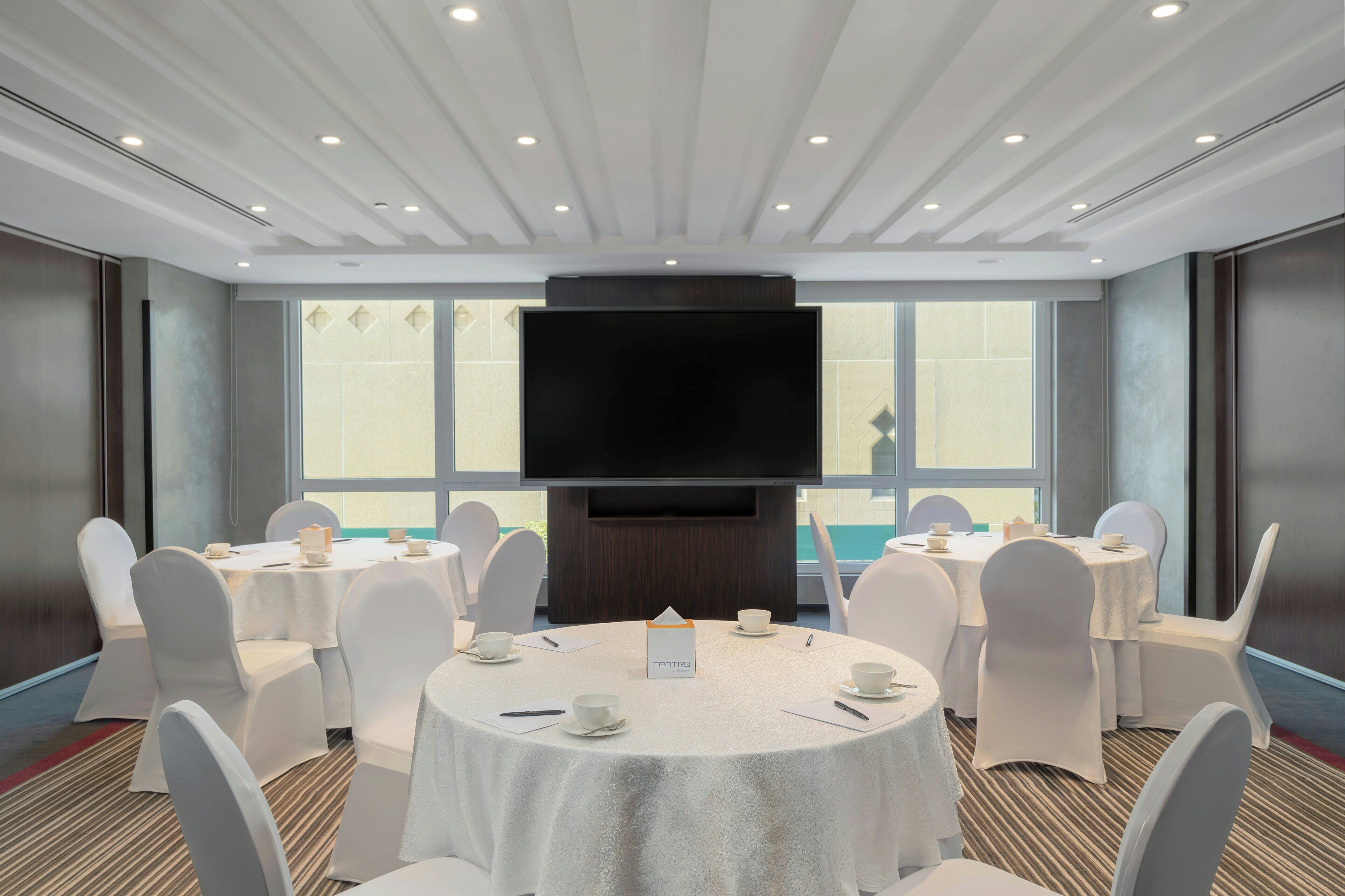 A conference room with round tables, white chairs, a large flat-screen TV, and a window with a view.