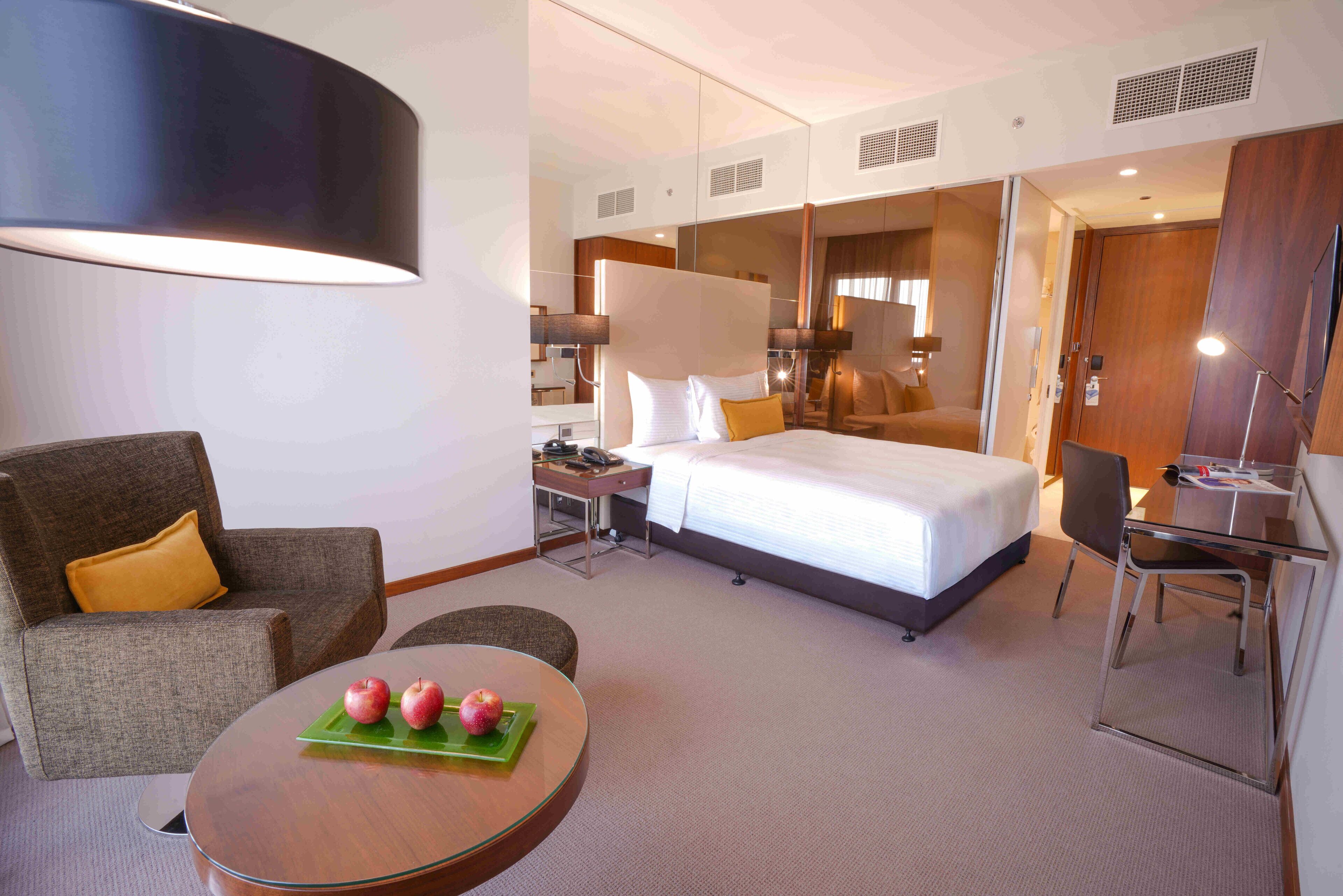 A hotel room with a bed, a desk, a chair, a sofa, and a table with apples.