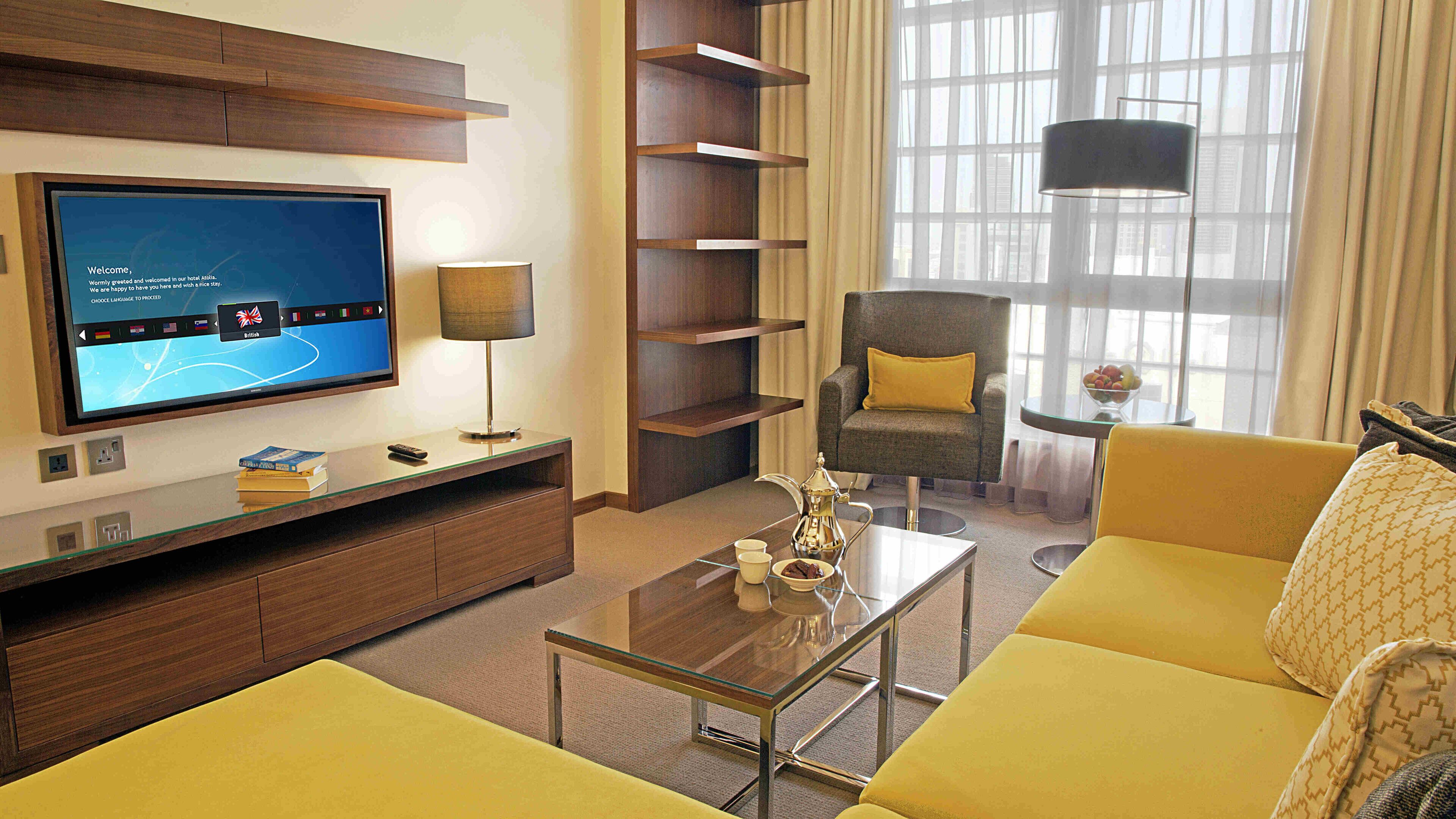 A modern hotel room with a flat-screen TV, a glass coffee table, a sofa, a chair, and a bookshelf.