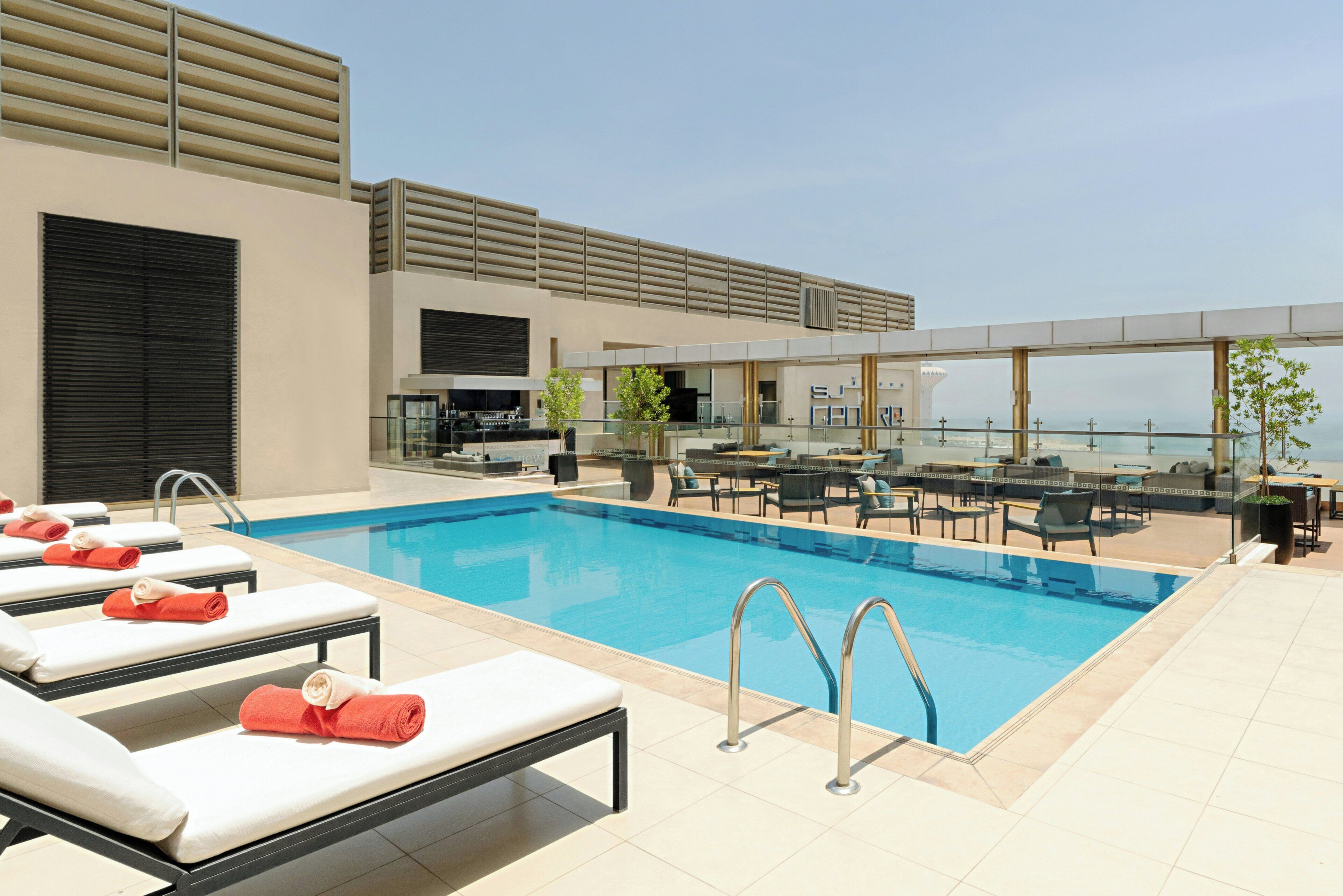 A rooftop pool with lounge chairs, a bar area, and a view of the city.