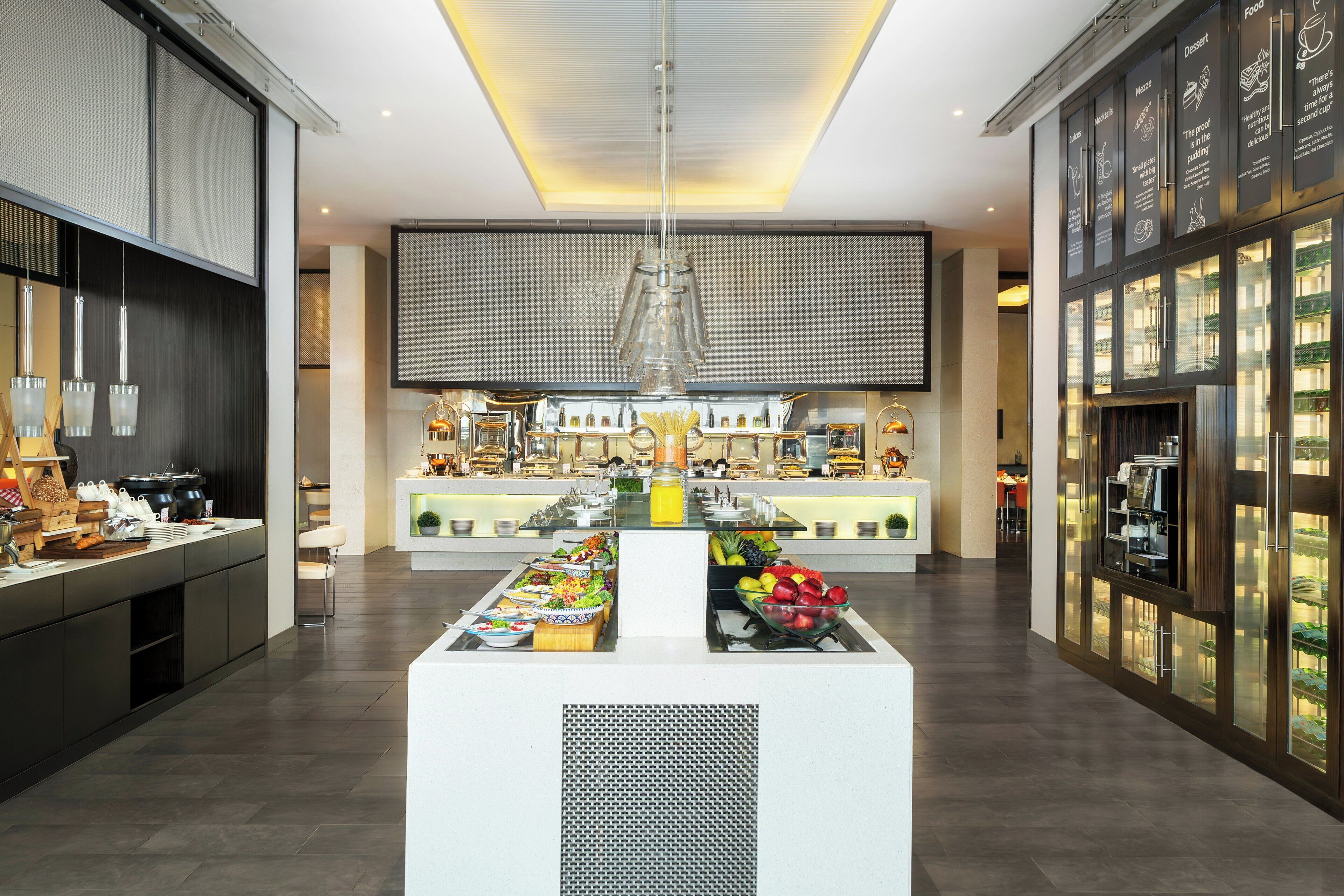 A modern hotel lobby with a central buffet, a large screen, and a well-lit reception area.