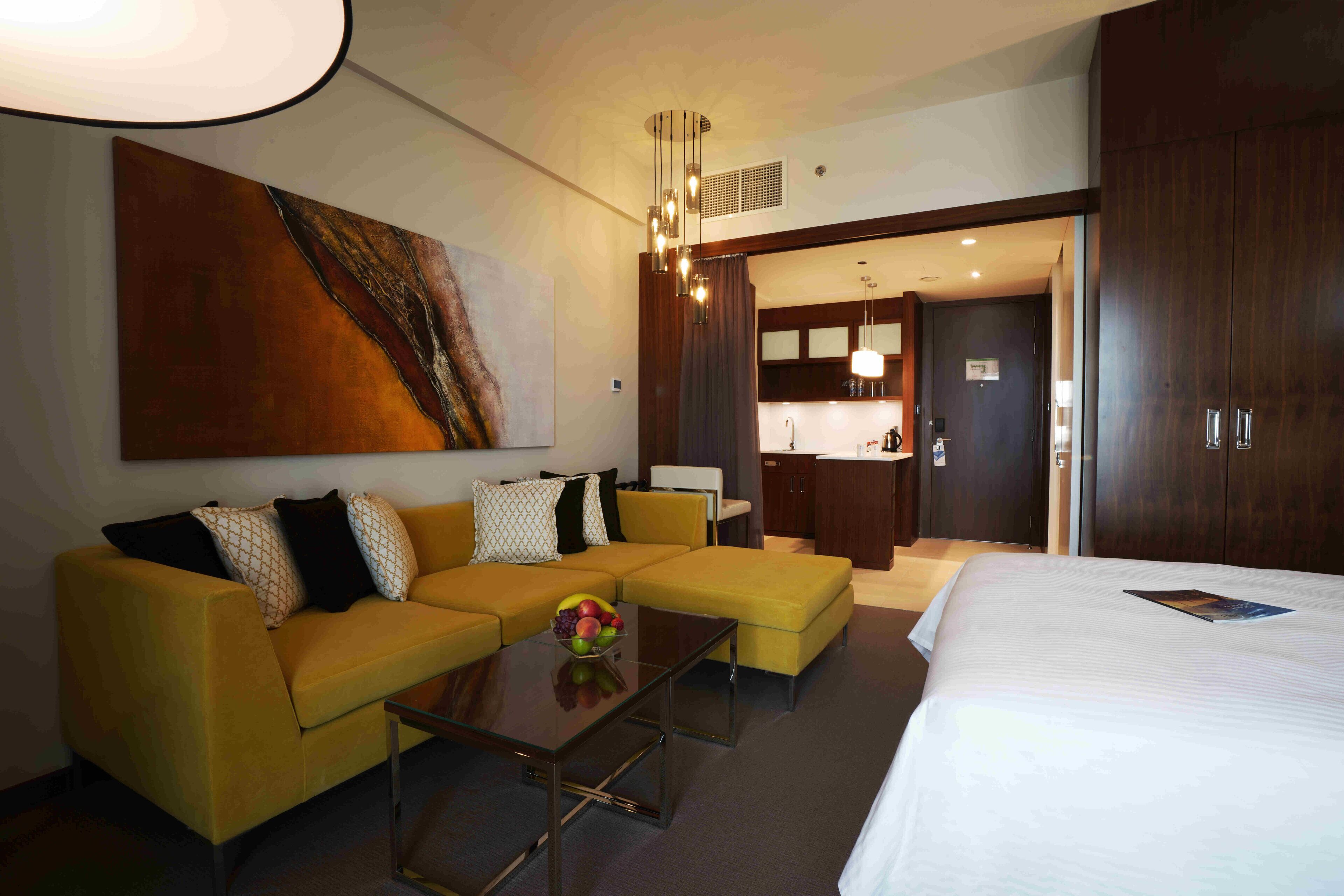A modern hotel room with a yellow sofa, a glass coffee table, a large bed, and a kitchen area visible through an open door.