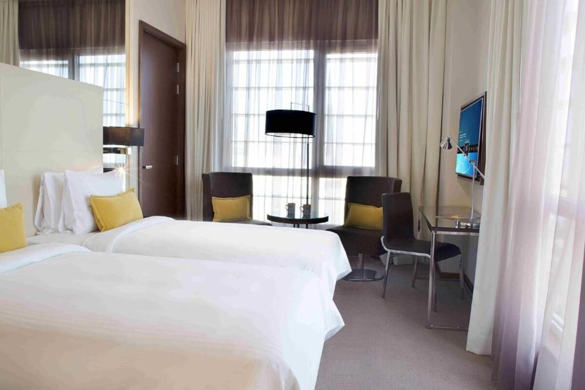 A hotel room with two beds, a desk, a chair, a TV, and a lamp.