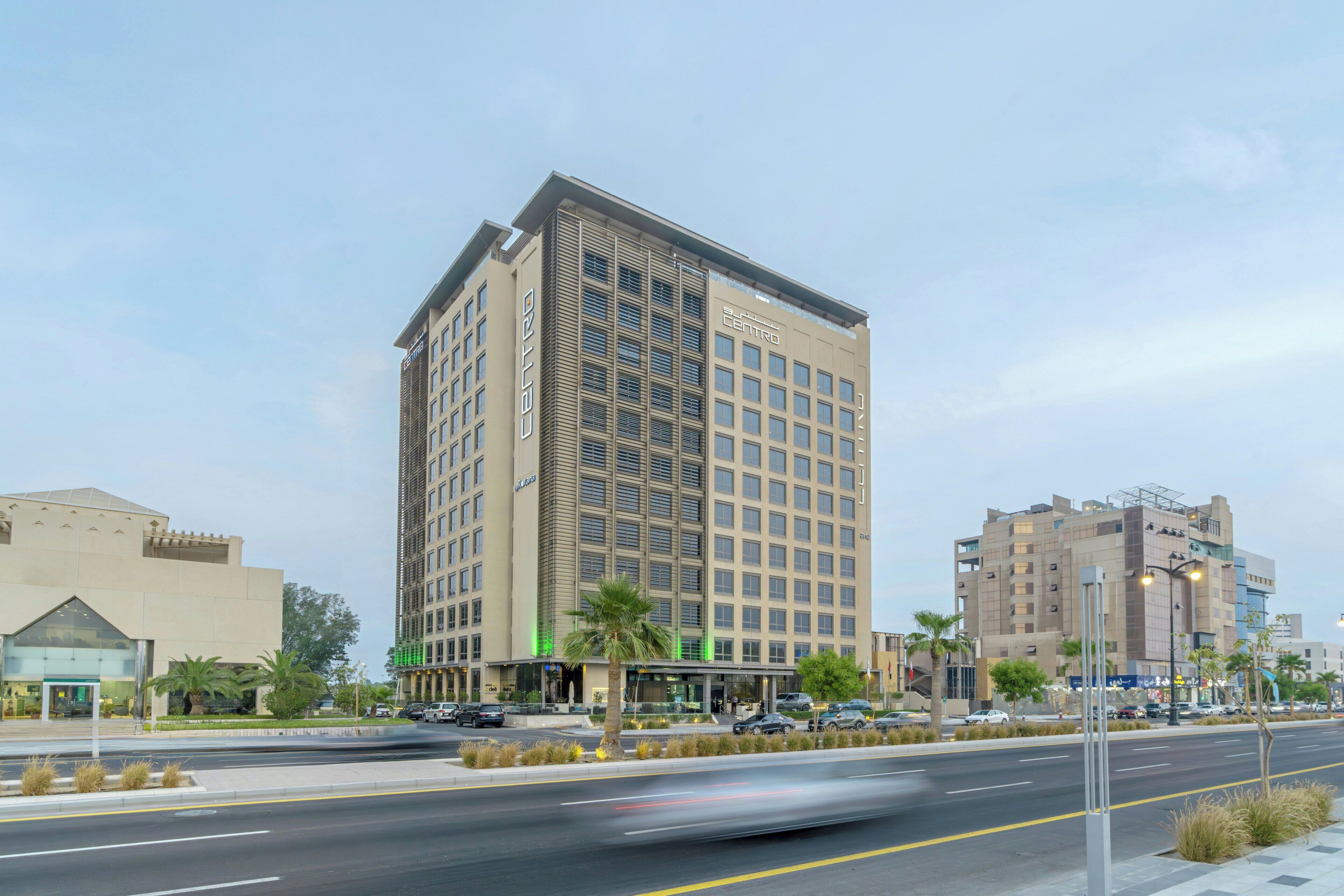 Un hôtel à plusieurs étages au design moderne, doté d’une entrée imposante, d’un espace de stationnement et d’une vue sur la rue, avec des palmiers et d’autres bâtiments.