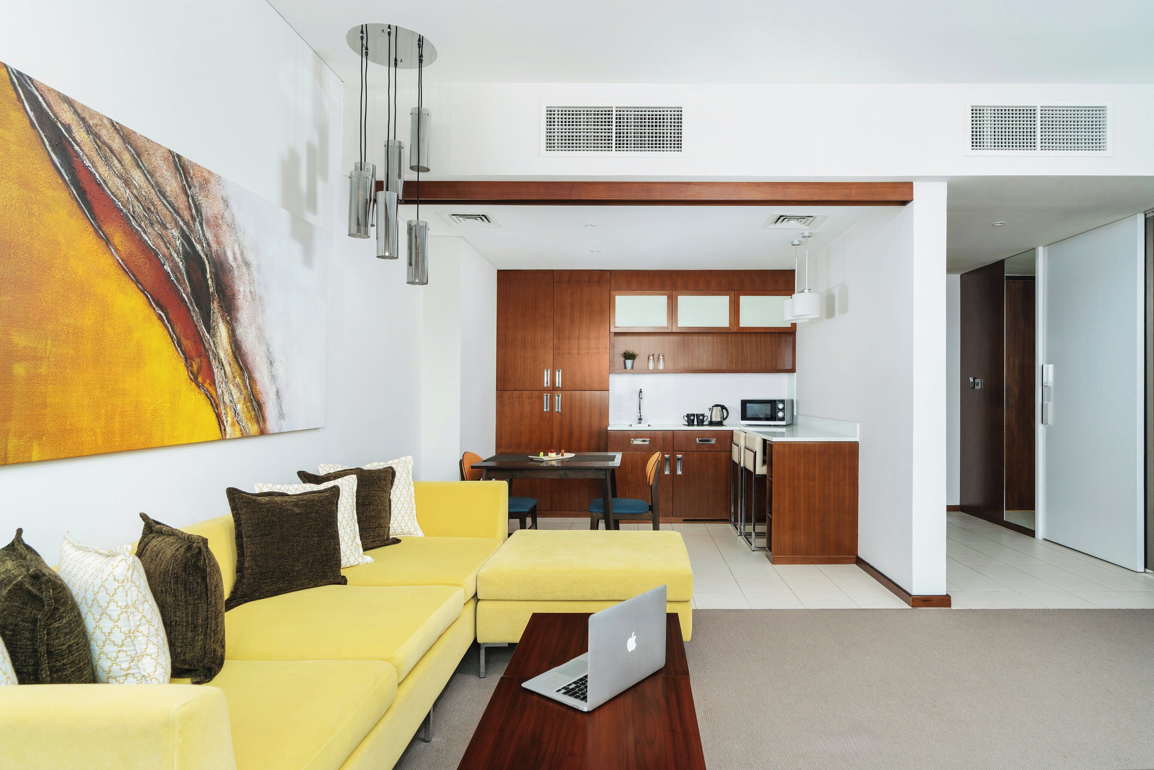A modern living room with a yellow sofa, a dining area, and a kitchen with wooden cabinets and white appliances.