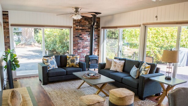 A cozy living room with a fireplace, two sofas, a coffee table, and a large window with a view of greenery. - Historic Creswell Farmhouse Pool+New Spa - 13 min to Eugene, U of O, & Wineries! (Creswell)