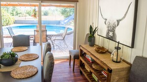 A dining area with a pool view, a framed artwork, and a wooden side table with books. - Historic Creswell Farmhouse Pool+New Spa - 13 min to Eugene, U of O, & Wineries! (Creswell)