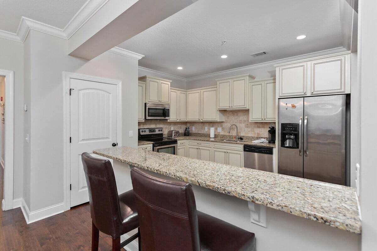 A modern kitchen with white cabinetry, granite countertops, stainless steel appliances, and a bar area with leather chairs.