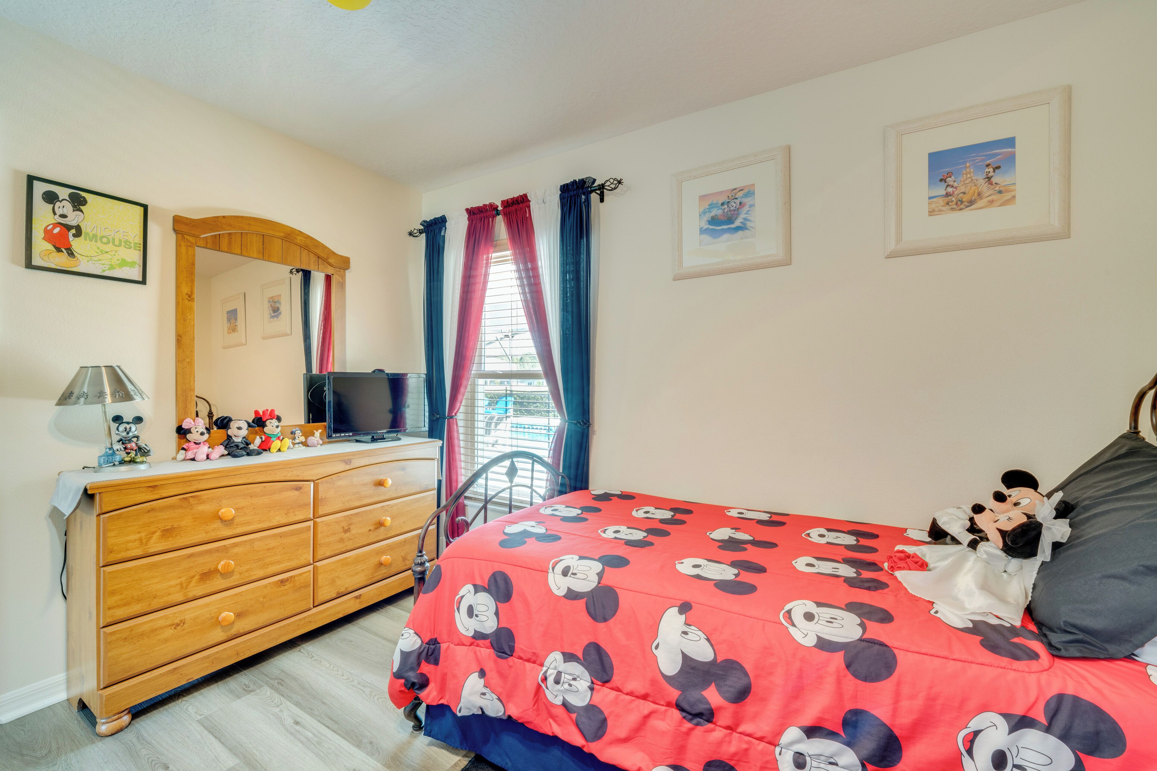 Une chambre avec une couvre-lit aux motifs de Mickey Mouse, une commode en bois, un téléviseur et une fenêtre avec des rideaux.