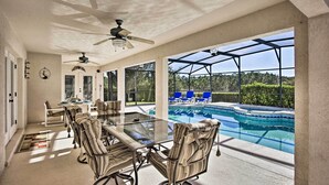 En overdækket terrasse med et bord i glas og stole, en ventilator i loftet og udsigt til en swimmingpool.