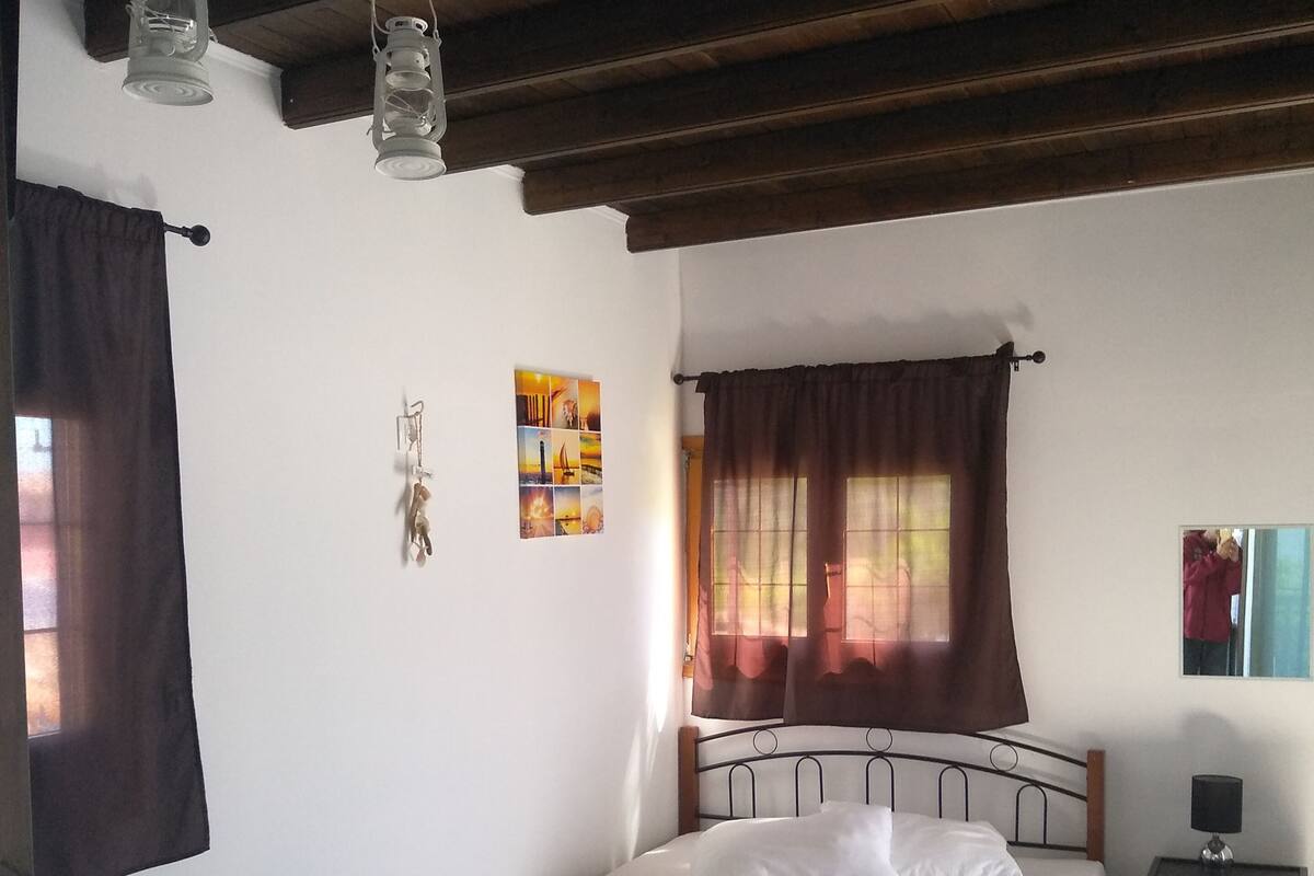 A bedroom with a single bed, a small table, a chair, a nightstand, and a window with curtains.
