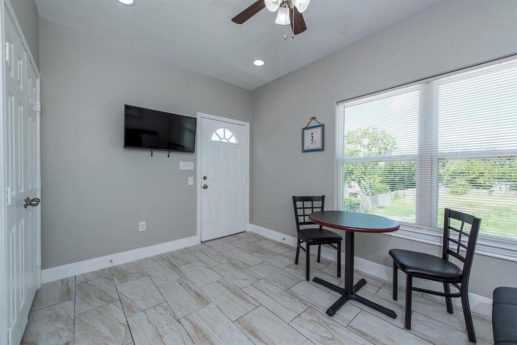 A room with a flat-screen TV mounted on the wall, a white door, a window with blinds, and a table with two chairs.