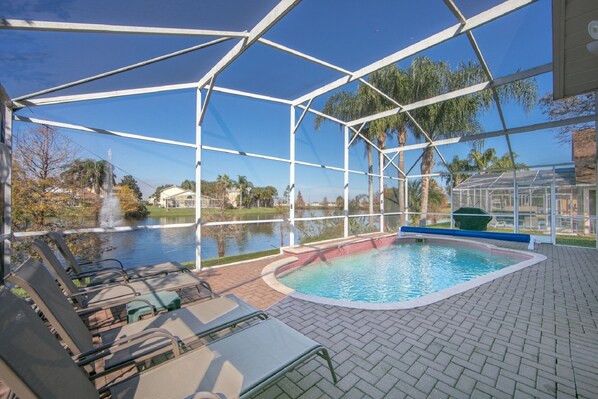 A covered outdoor swimming pool with lounge chairs and a view of a lake and palm trees.