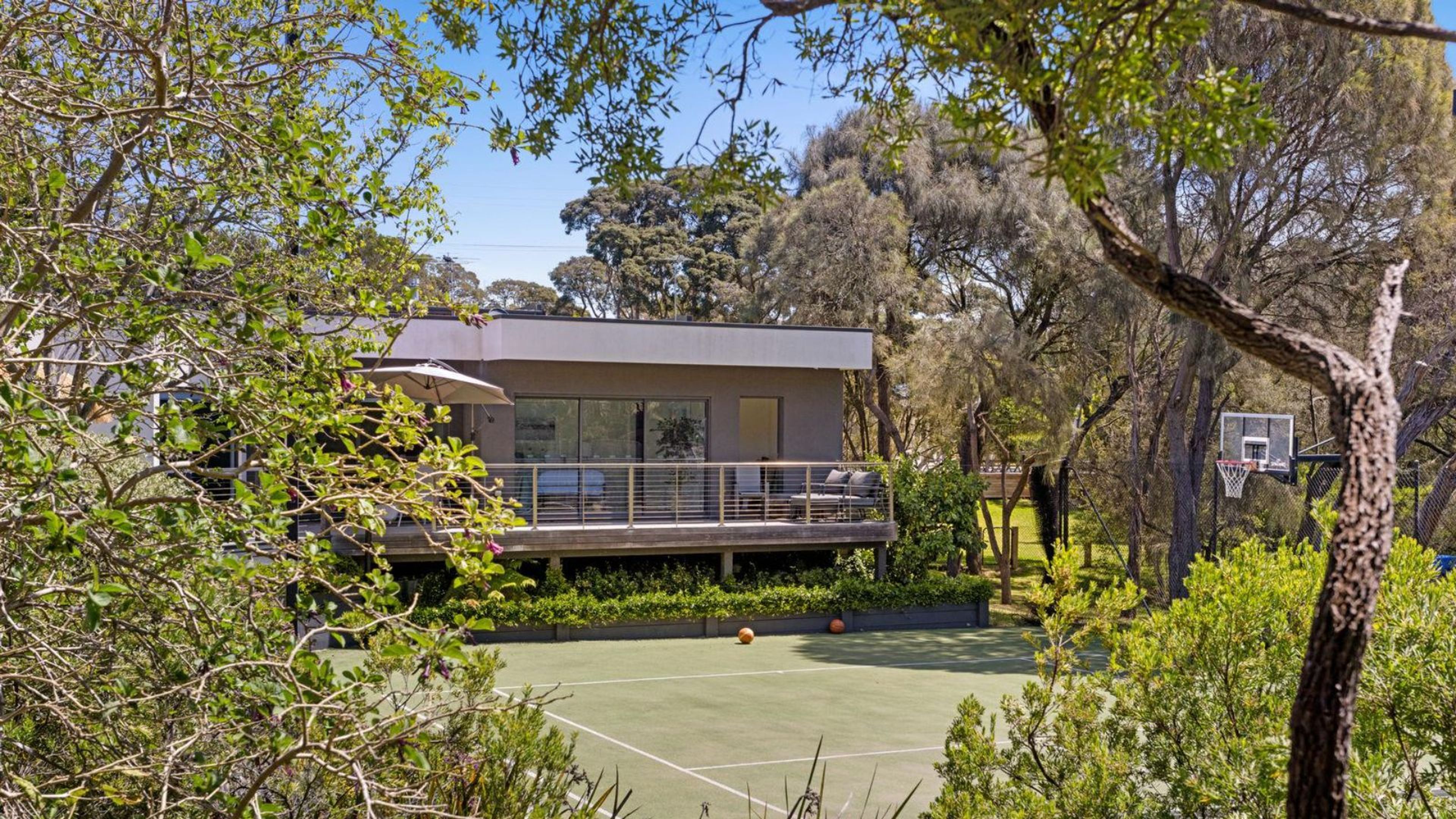 Una casa moderna con balcón que da a una cancha de tenis, rodeada de abundante vegetación.