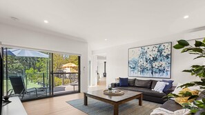 A modern living room with a glass door leading to an outdoor area, a large abstract painting on the wall, and a leather sofa. - Coastal Hideaway in Blairgowrie (BLAIRGOWRIE)
