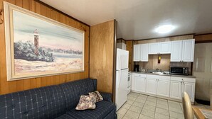 Un salon avec un canapé et un tableau, une cuisine avec des meubles blancs et un four à micro-ondes, et un sol carrelé.